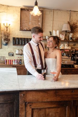 A bride and groom share a romantic moment at a rustic bar, holding drinks and smiling at each other on their wedding day.