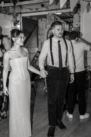 Black and white photo of a smiling bride in a corset gown holding hands with a groom in suspenders at a wedding reception.