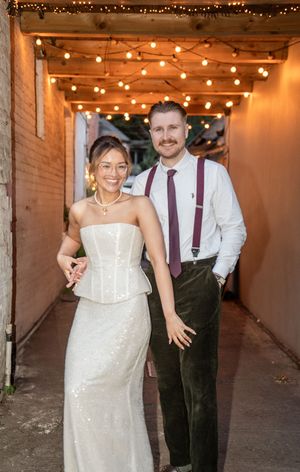 Bride and groom posing under string lights in a romantic alley, she in a white sequin gown, he in suspenders and tie.