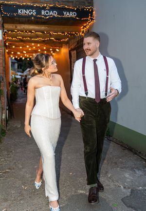 Couple holding hands walking outside Kings Road Yard venue, bride in white corset dress, groom in suspenders.