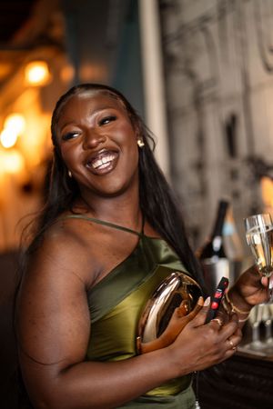 A joyful woman in a green satin dress holds a gold clutch and champagne glass at an elegant evening event.