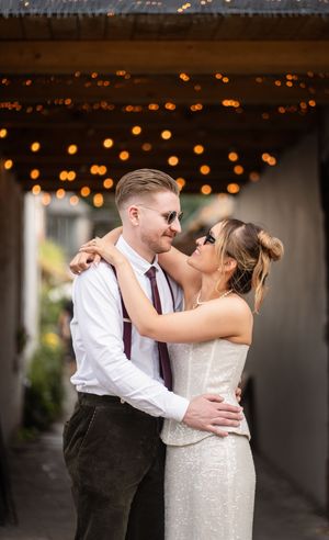 Couple in wedding attire embrace under warm string lights, groom in white shirt with burgundy tie and suspenders.