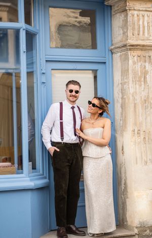 A stylish couple poses by a blue door, man in suspenders and woman in a white strapless dress with sunglasses.