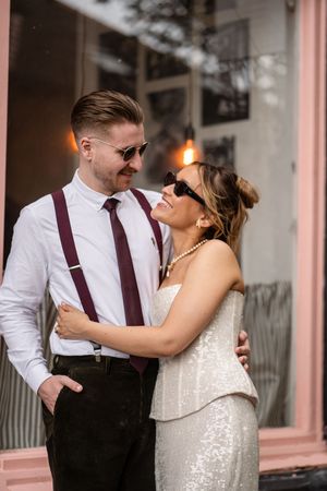 Stylish bride and groom in sunglasses embracing outside a pink building, she in sequin dress, he in white shirt with suspenders.