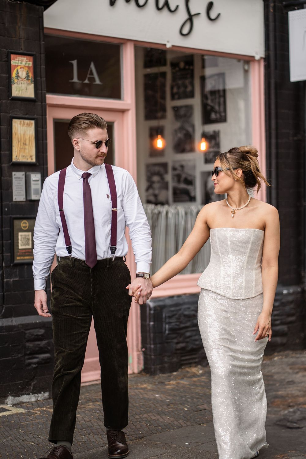 Bride and groom holding hands walking past a pink storefront, wearing sunglasses and wedding attire on their special day.