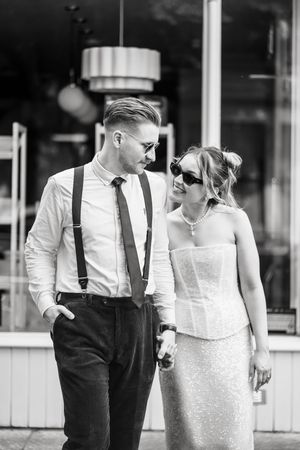 Black and white photo of stylish couple in wedding attire, man in suspenders and tie, woman in strapless dress with sunglasses.