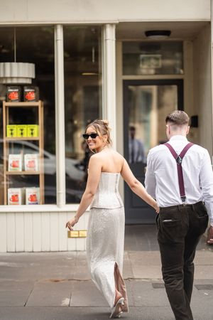 Bride in white strapless gown and groom in suspenders walk hand in hand along a city street.