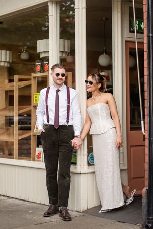 Wedding couple holding hands outside a shop, groom in suspenders and bride in white strapless gown with sunglasses.