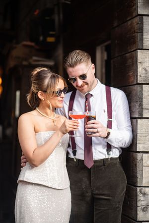A couple in wedding attire clinks cocktail glasses outdoors, man wearing suspenders and tie, woman in sequined dress.