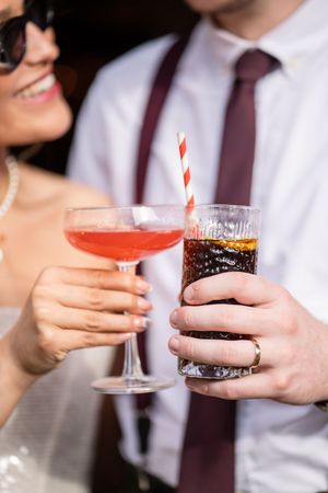 Bride and groom clinking cocktail glasses, she holds pink drink, he holds cola with straw.