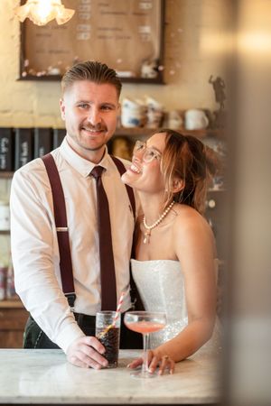Smiling bride and groom holding drinks at a bar counter, bride wearing strapless gown and pearl necklace.