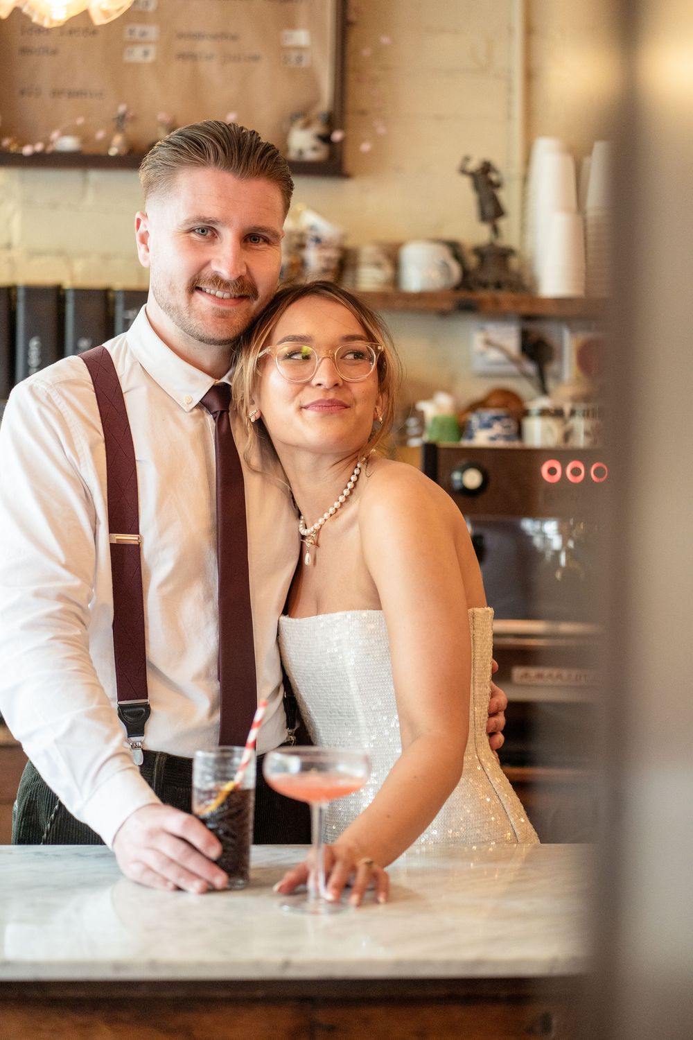 Smiling bride and groom pose together at a cafe bar, holding cocktails, dressed in wedding attire with suspenders and white gown.