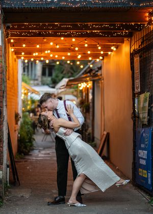 Couple sharing a romantic dip kiss under string lights in a charming alley, groom in white shirt, bride in silver dress.