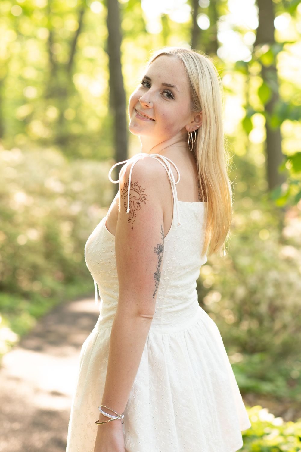 Blonde woman in white sundress with tattoos smiling over shoulder on sunny forest path.