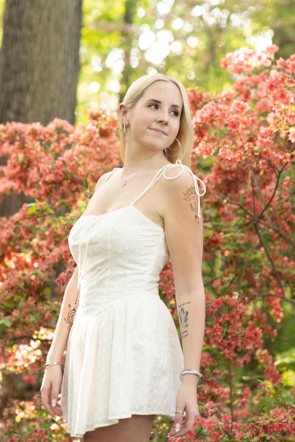 Young blonde woman with tattoos in white sundress posing outdoors among vibrant pink azalea bushes in a garden.