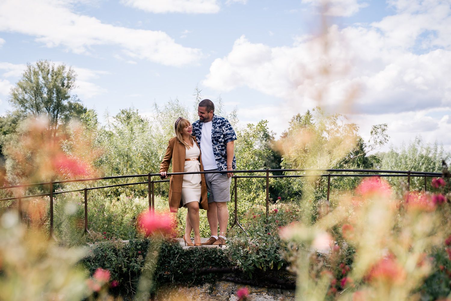 Couple standing on a garden bridge surrounded by blooming red flowers and lush greenery under a cloudy sky.