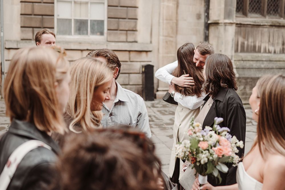 Wedding guests embrace and celebrate outdoors near a historic stone building, with a bride holding a pastel floral bouquet.