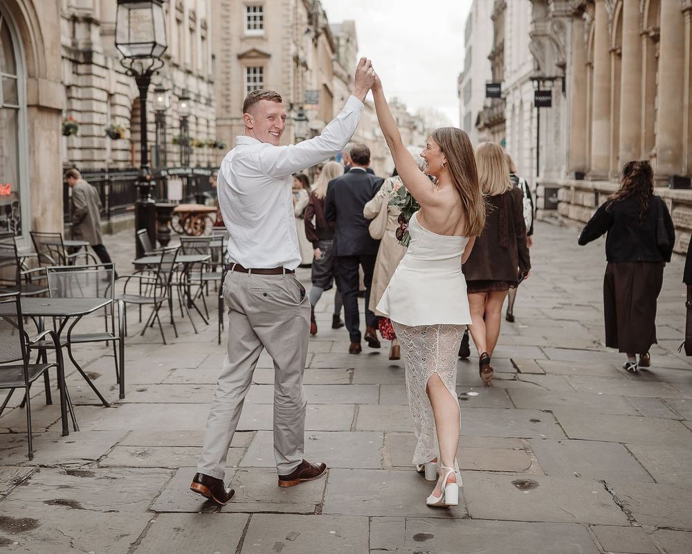 Couple dancing joyfully on a cobblestone street in London, woman in white dress, man in white shirt and gray trousers.