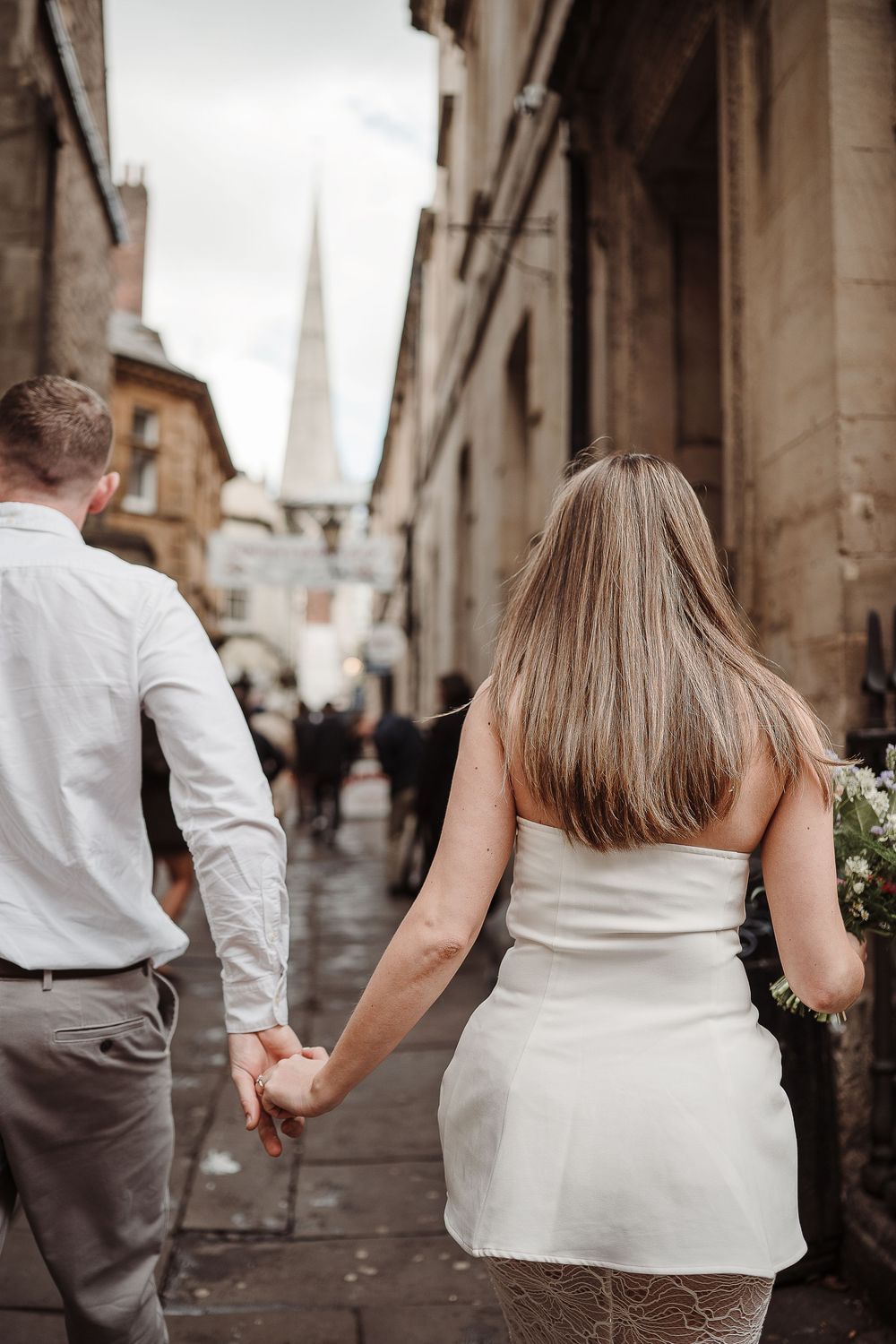 Newlywed couple walking hand in hand through a narrow cobblestone street with a church spire in background.