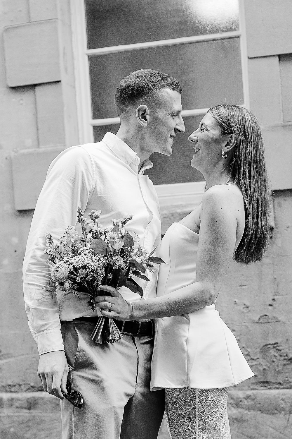 Black and white photo of a smiling couple in wedding attire sharing a romantic moment while holding a bouquet.