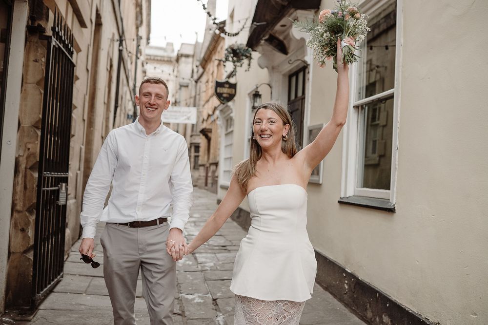 Joyful newlyweds walk cobblestone street, bride in white strapless gown raises bouquet, groom in white shirt holds her hand.