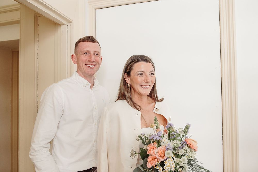 Smiling couple in white attire posing indoors, bride holding a pastel bouquet of peach and white flowers.