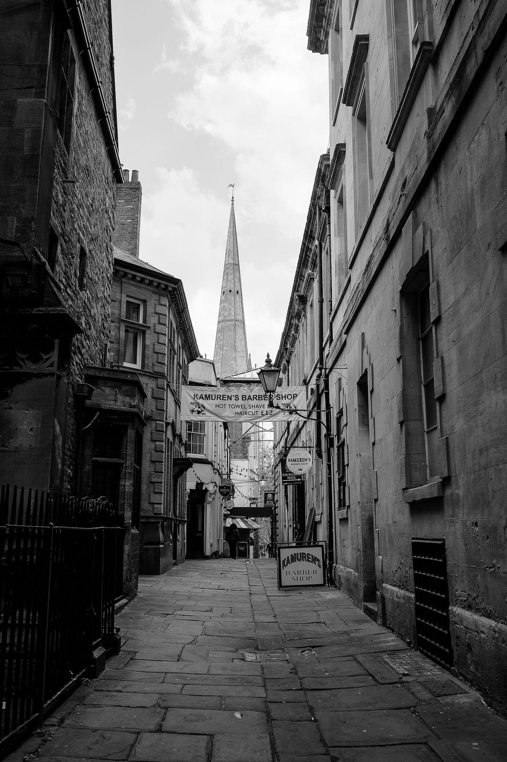 Black and white photo of a narrow historic alley with a church spire and barber shop signs visible.