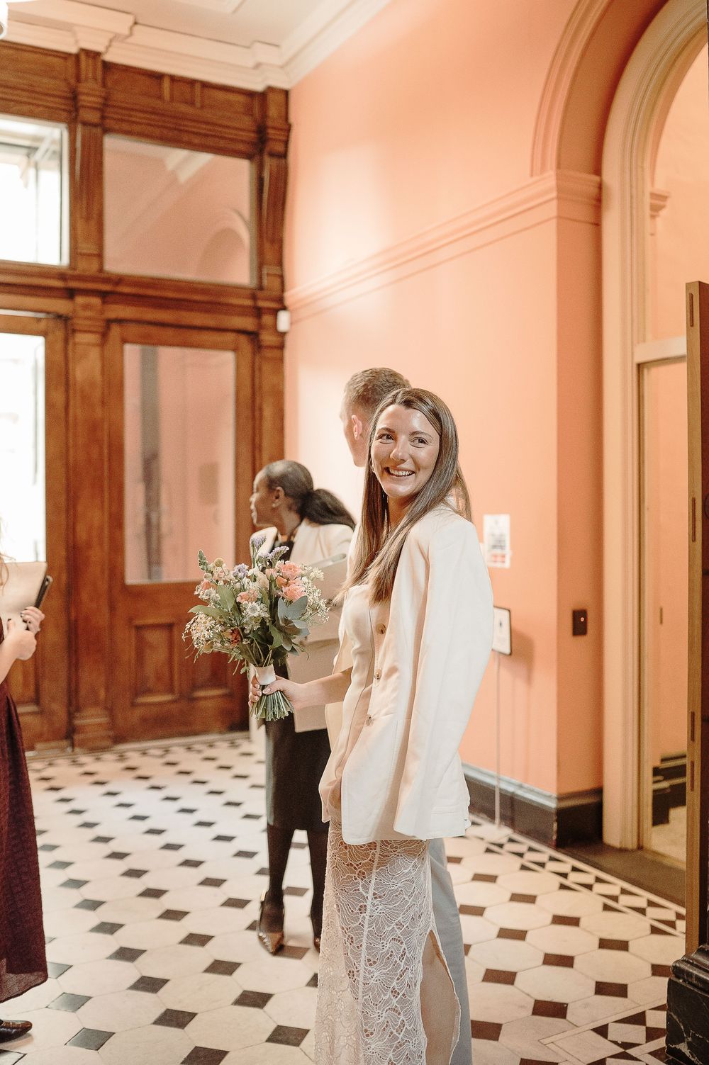 Smiling bride in white lace dress and blazer holding a floral bouquet in an elegant pink-walled venue.