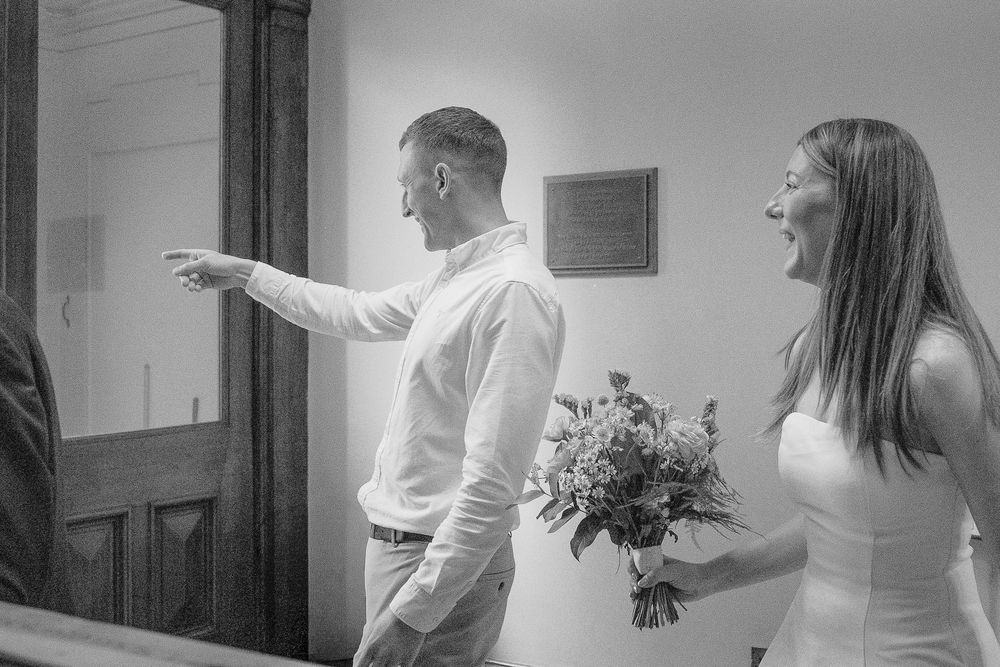 Black and white photo of a bride holding bouquet laughing with groom pointing during wedding ceremony.