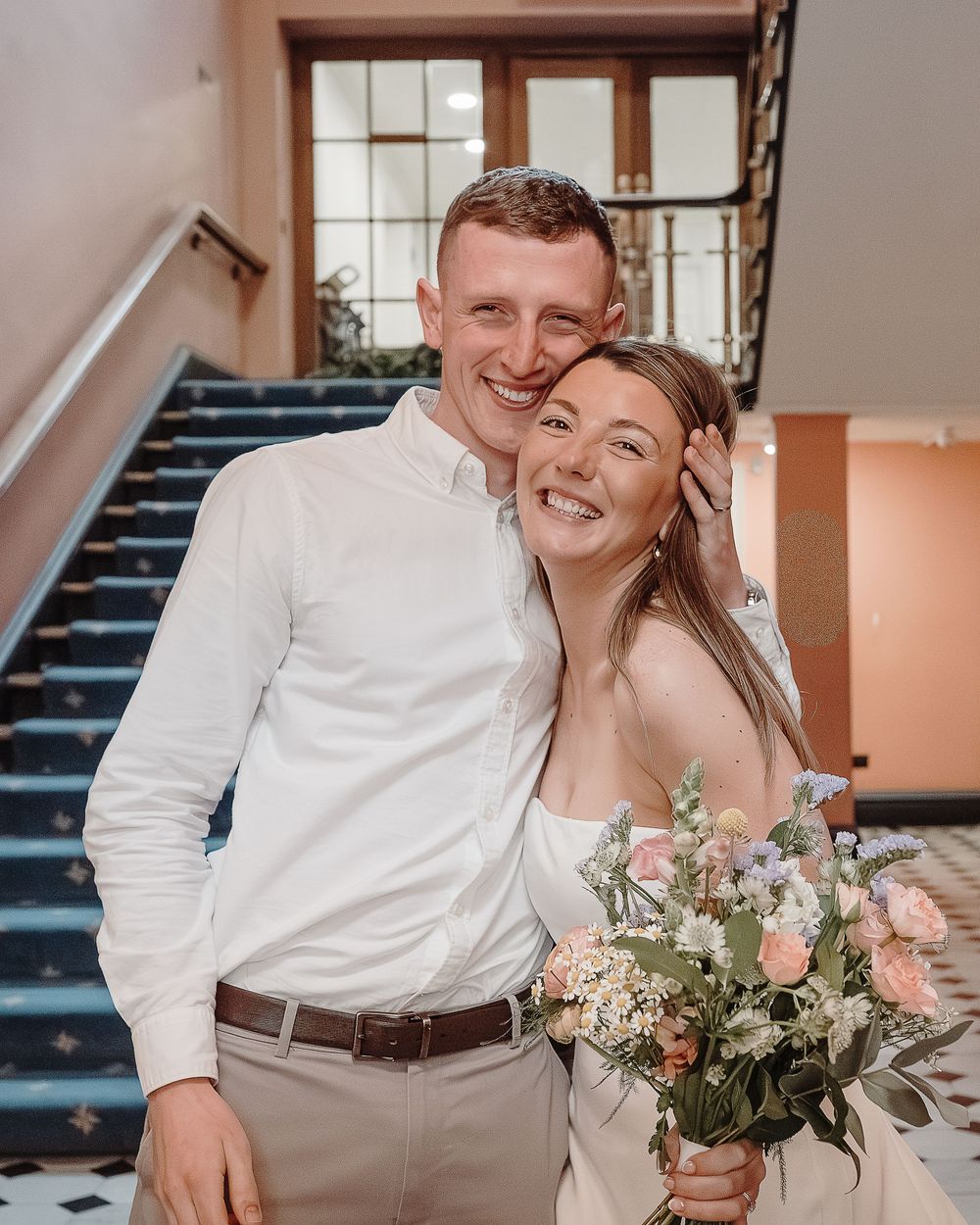 Smiling newlywed couple posing on staircase, bride holding pastel floral bouquet in elegant white dress.