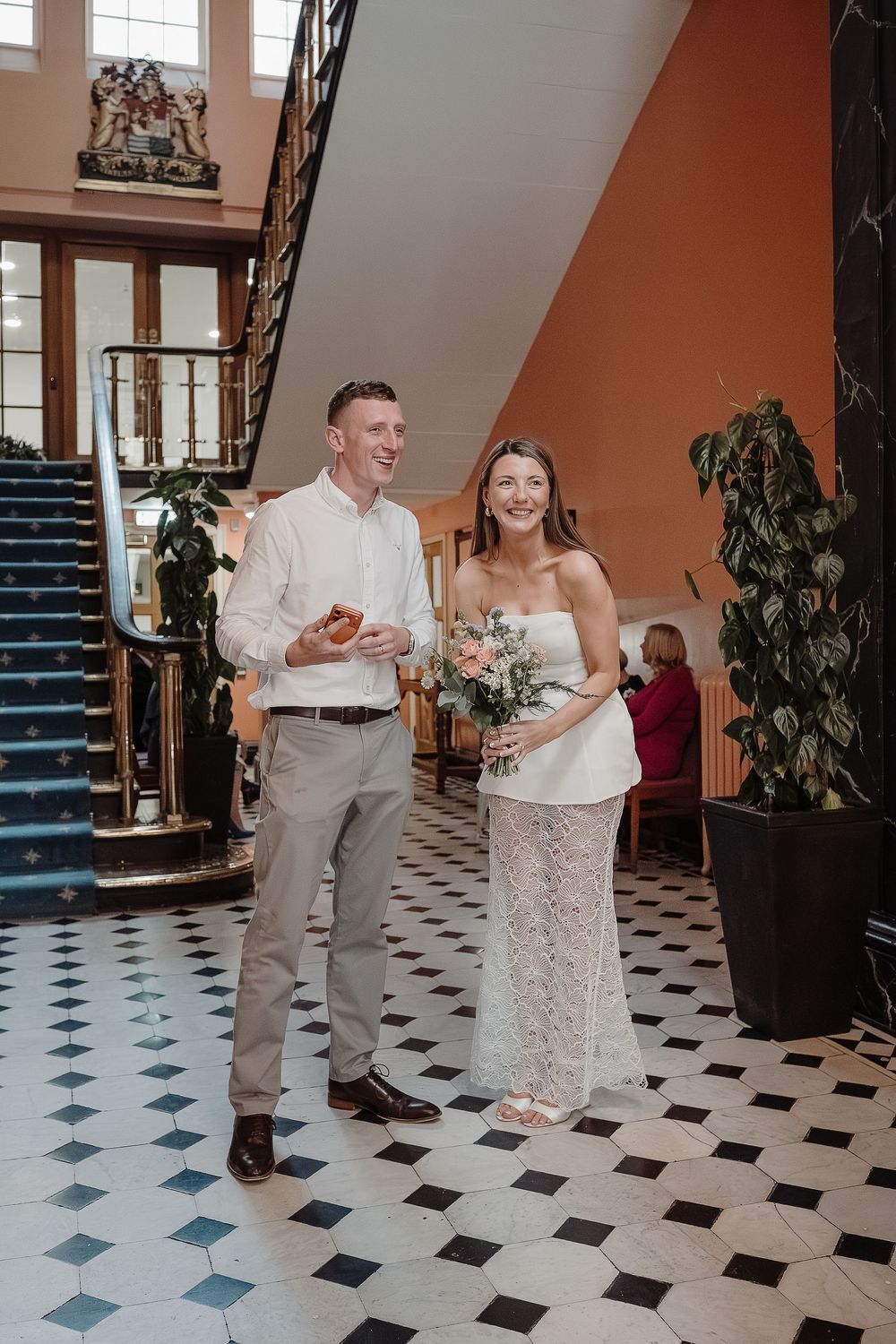 Happy couple laughing in elegant building lobby, bride holding floral bouquet in white strapless gown, groom in white shirt.