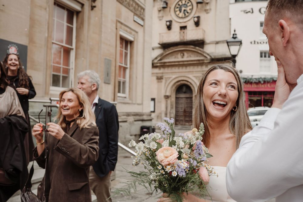 Joyful bride laughing outdoors holding a pastel bouquet, with guests celebrating after a wedding ceremony.