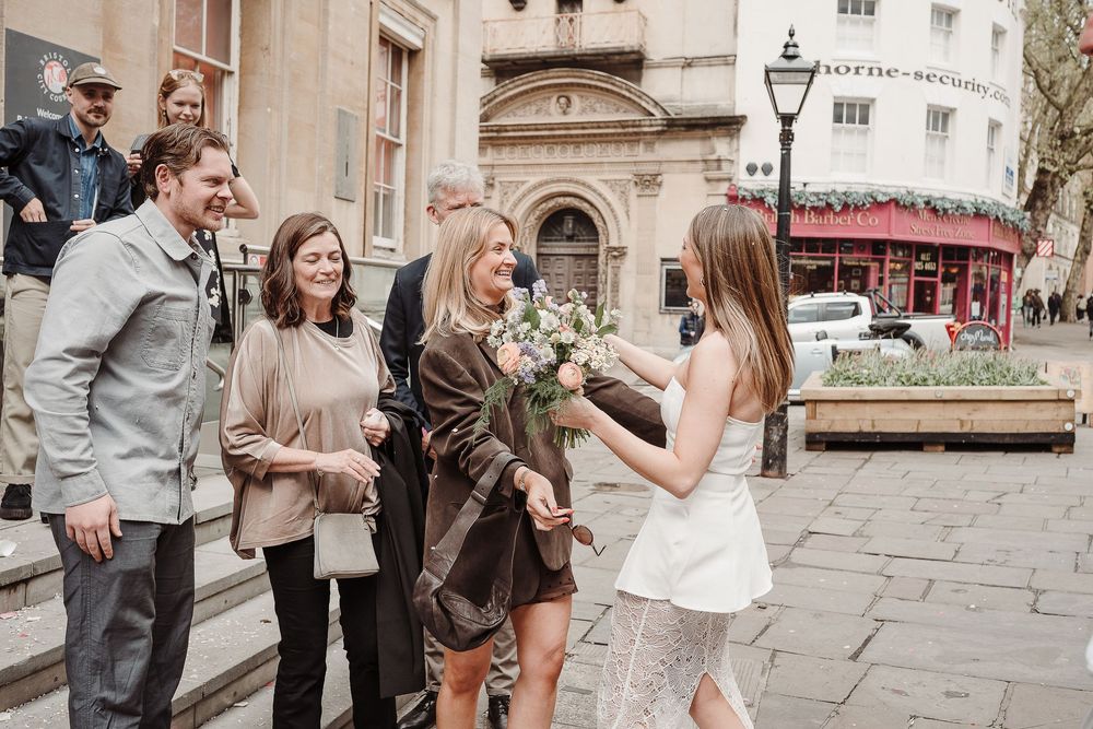 Bride in white dress receives bouquet from guest outside elegant building during intimate wedding celebration.