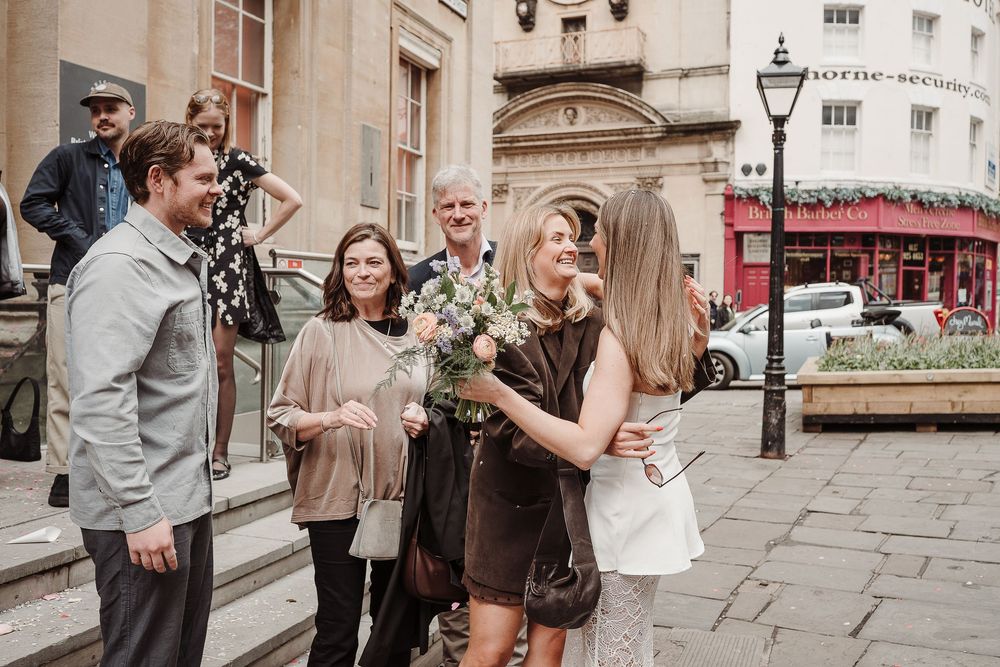 Wedding guests greeting bride outdoors on city street, sharing joyful embraces after ceremony.