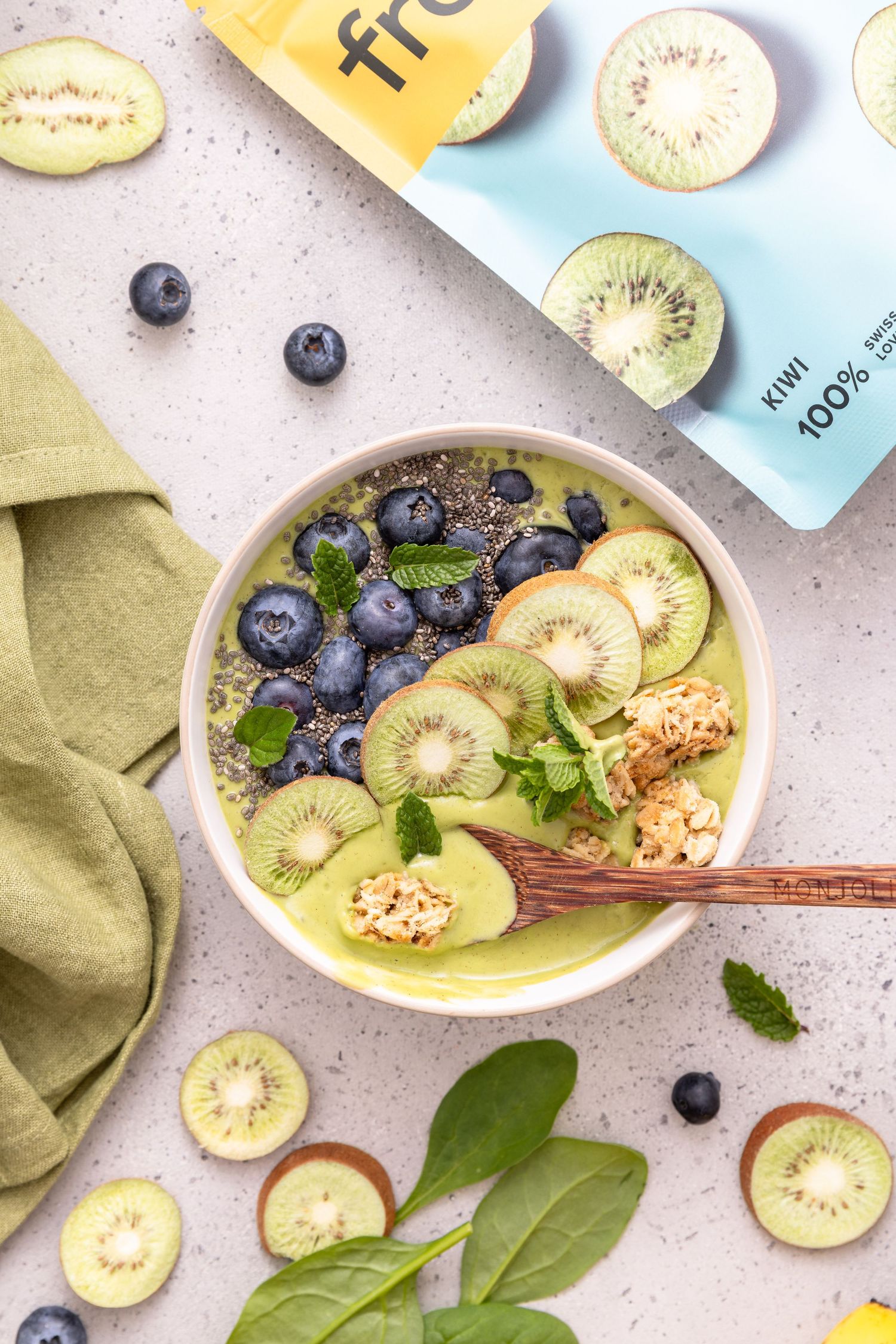Green smoothie bowl topped with kiwi slices, blueberries, chia seeds, and granola, with a kiwi juice pack nearby.