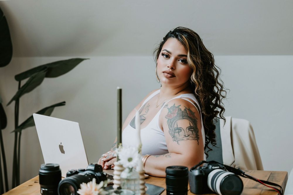 Tattooed female photographer sitting at desk with multiple camera lenses and Apple laptop in bright studio workspace.