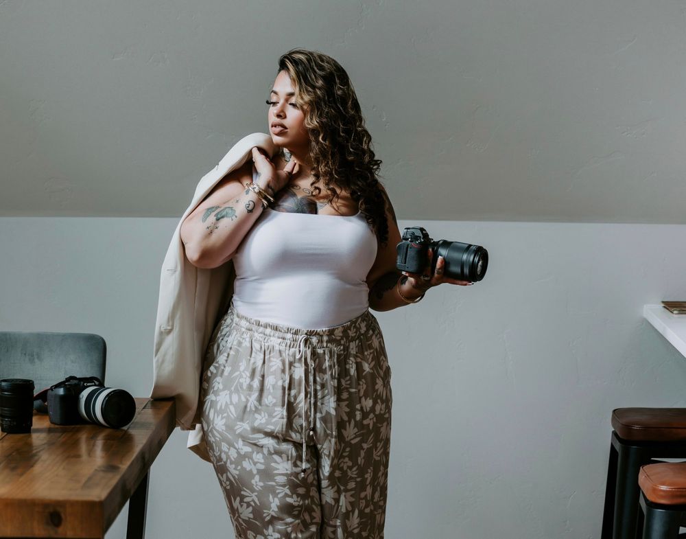 A plus-size woman photographer holds a Canon camera while posing confidently in a white bodysuit and floral skirt.