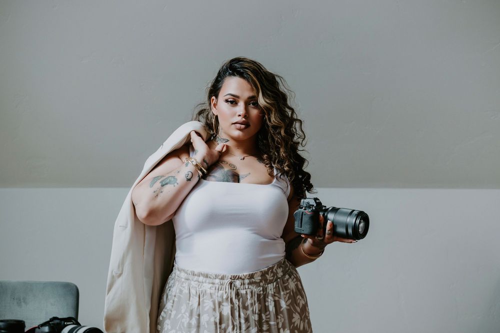Woman with curly hair holding a camera, wearing a white top and floral skirt, posing confidently indoors.
