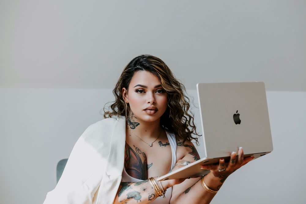 Tattooed woman with curly hair holding a MacBook laptop, wearing a white blazer, posing confidently indoors.