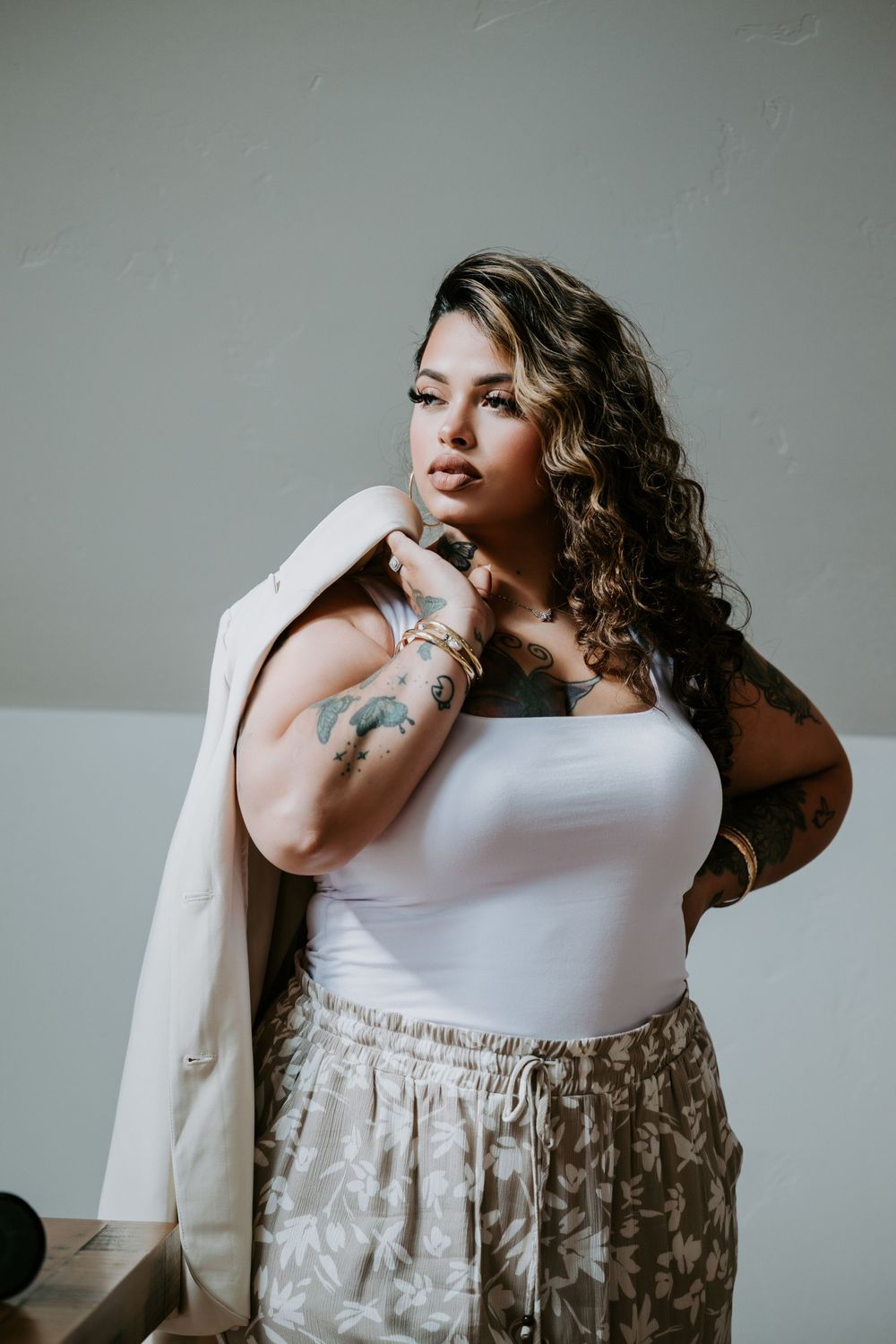 Plus-size woman with tattoos and curly hair posing in white bodysuit, floral skirt, and beige blazer.