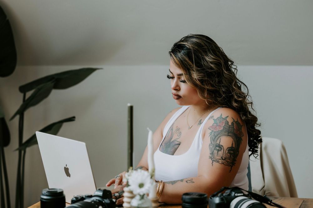 Woman with tattoos working on a MacBook laptop at a desk with cameras and flowers in a bright studio.