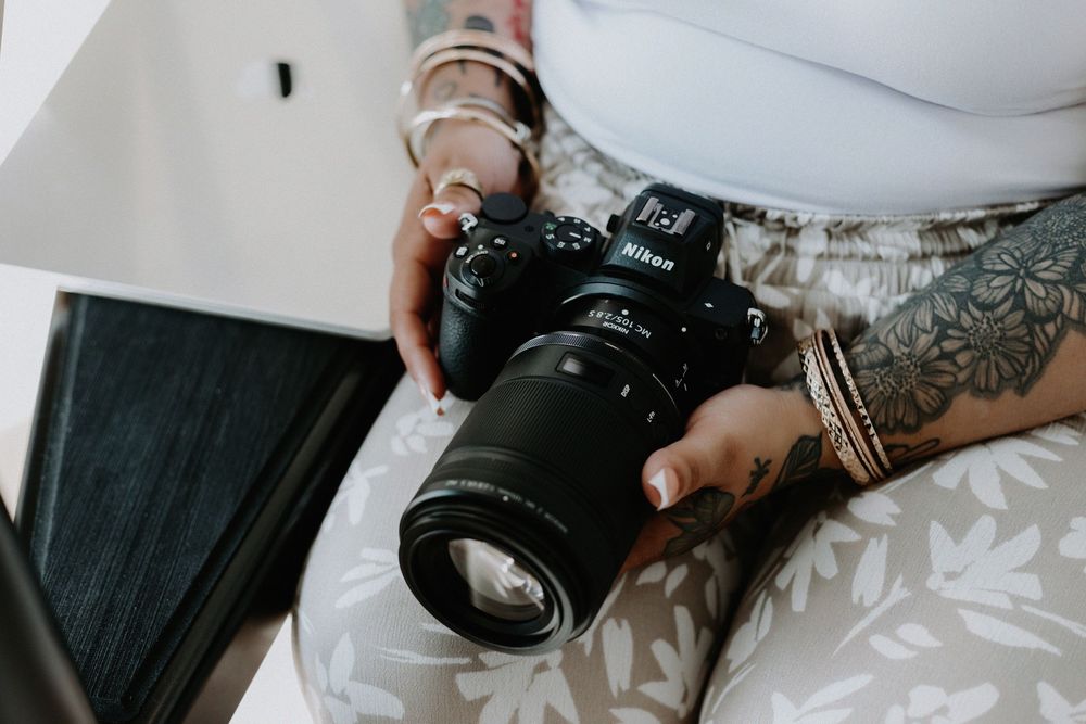 Person with tattoos and bracelets holding a Nikon DSLR camera with a large lens while seated.