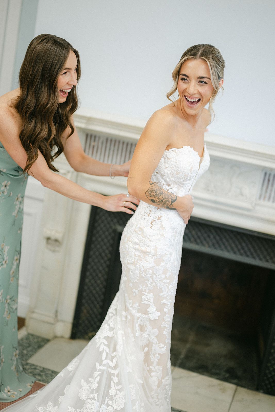 Armour House Wedding - A bride in a lace strapless gown laughs joyfully as her bridesmaid in sage green helps fasten her dress.