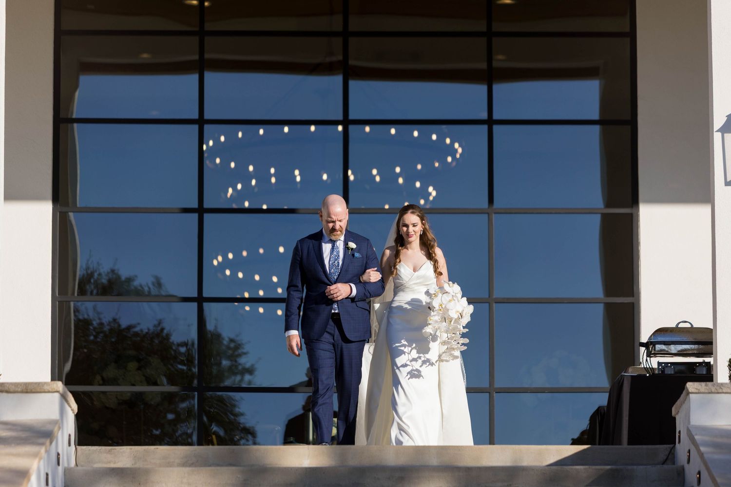 Bride and groom descend steps outside modern venue with large glass windows reflecting string lights at wedding ceremony.