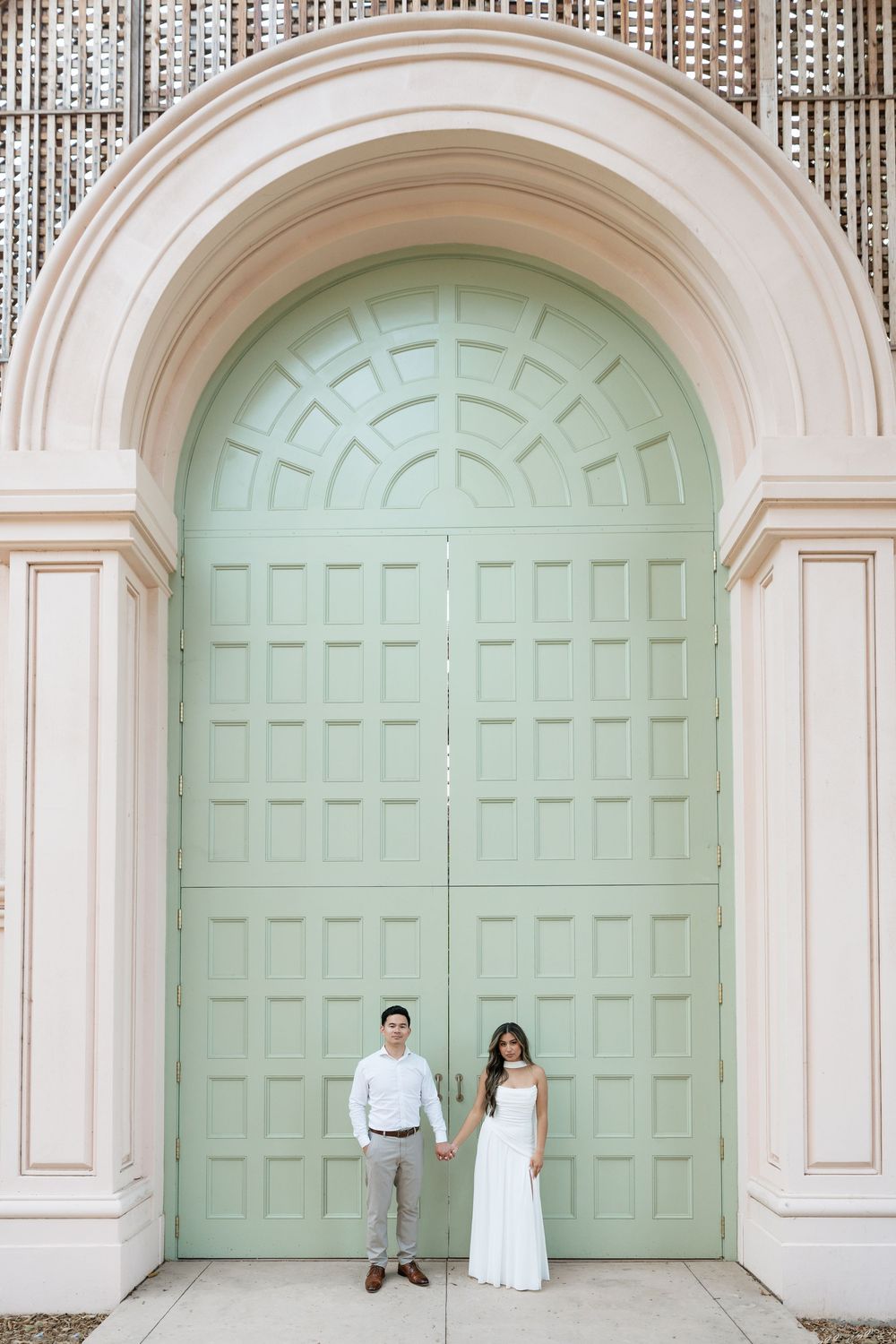 Engaged couple holding hands before large mint green arched doors of elegant building in white outfits.
