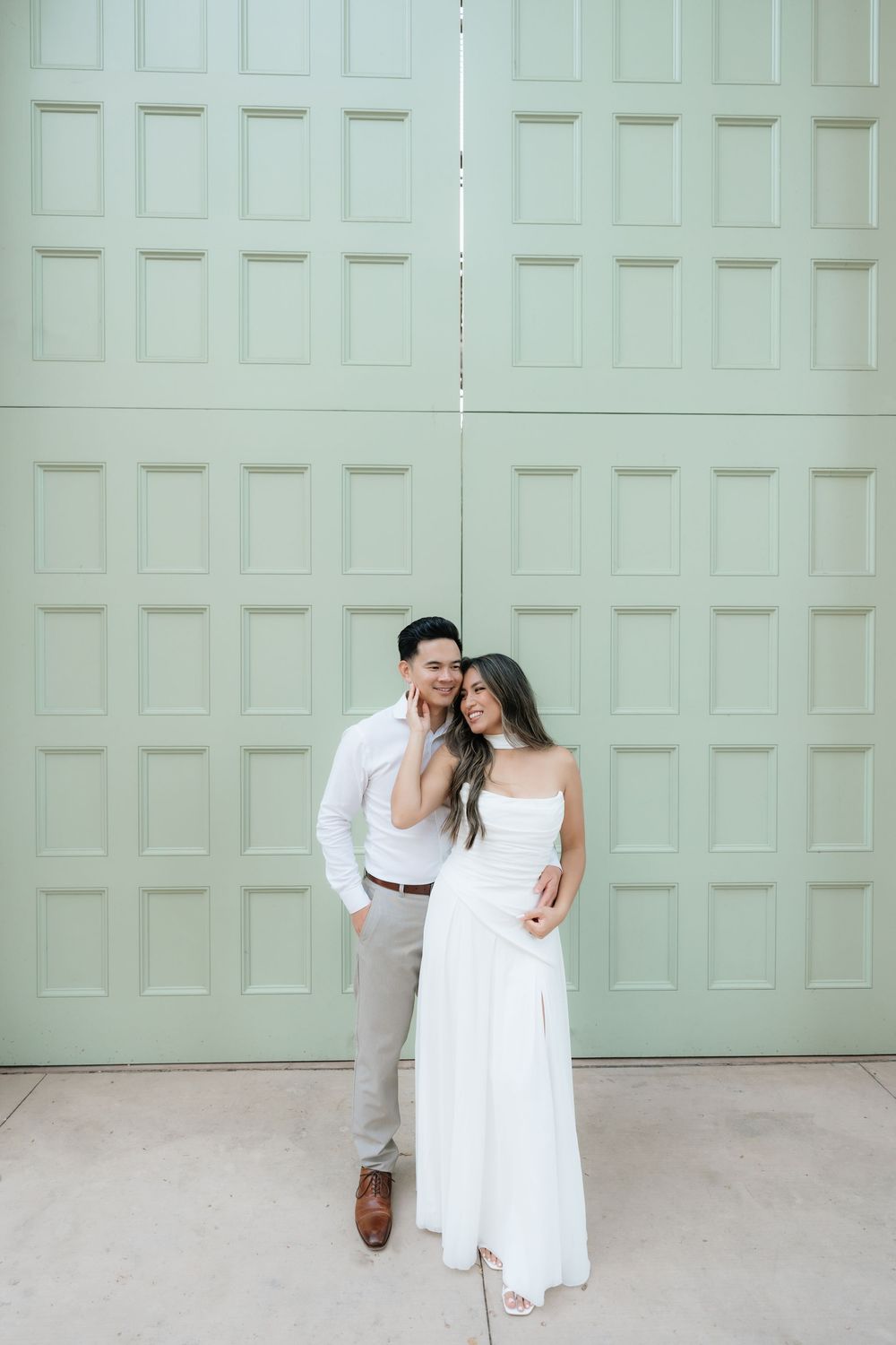 Smiling couple posing together in white outfits against a large mint green door backdrop.