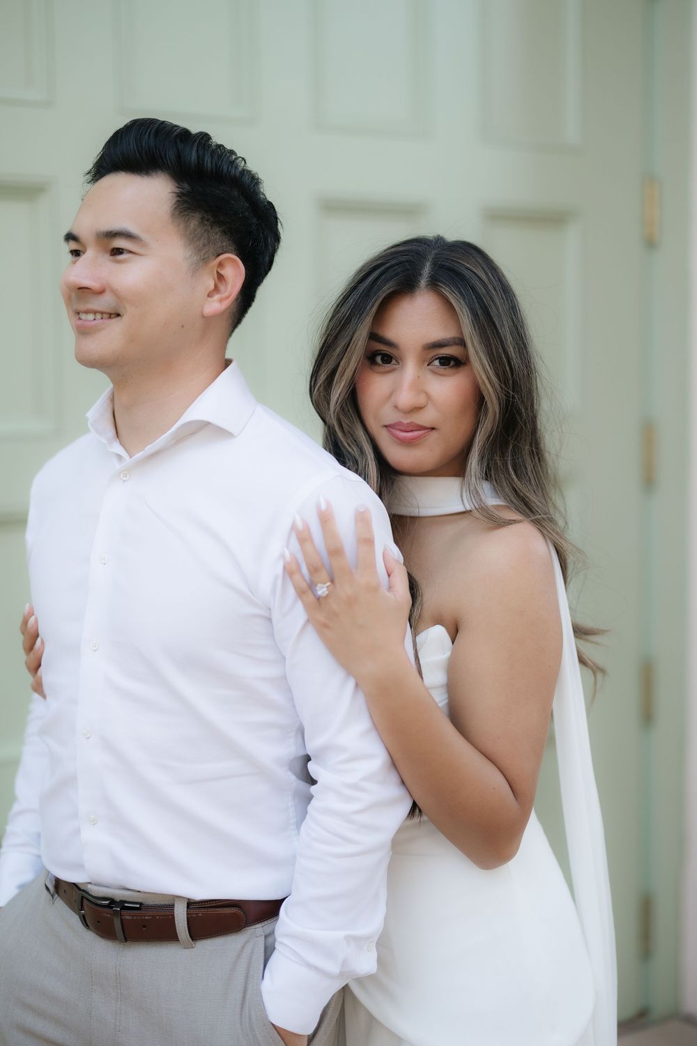 Bride in white halter gown embraces groom from behind showing engagement ring near a sage green door.
