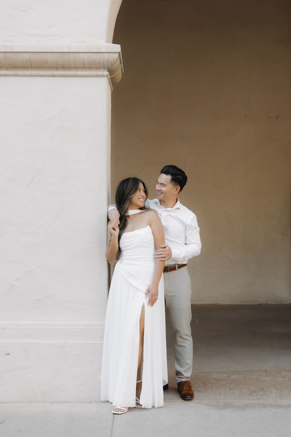 Couple in white wedding attire posing elegantly against a beige wall under an archway.