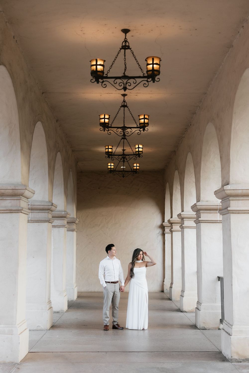 Couple holds hands under ornate chandeliers in an arched colonnade corridor at Balboa Park.