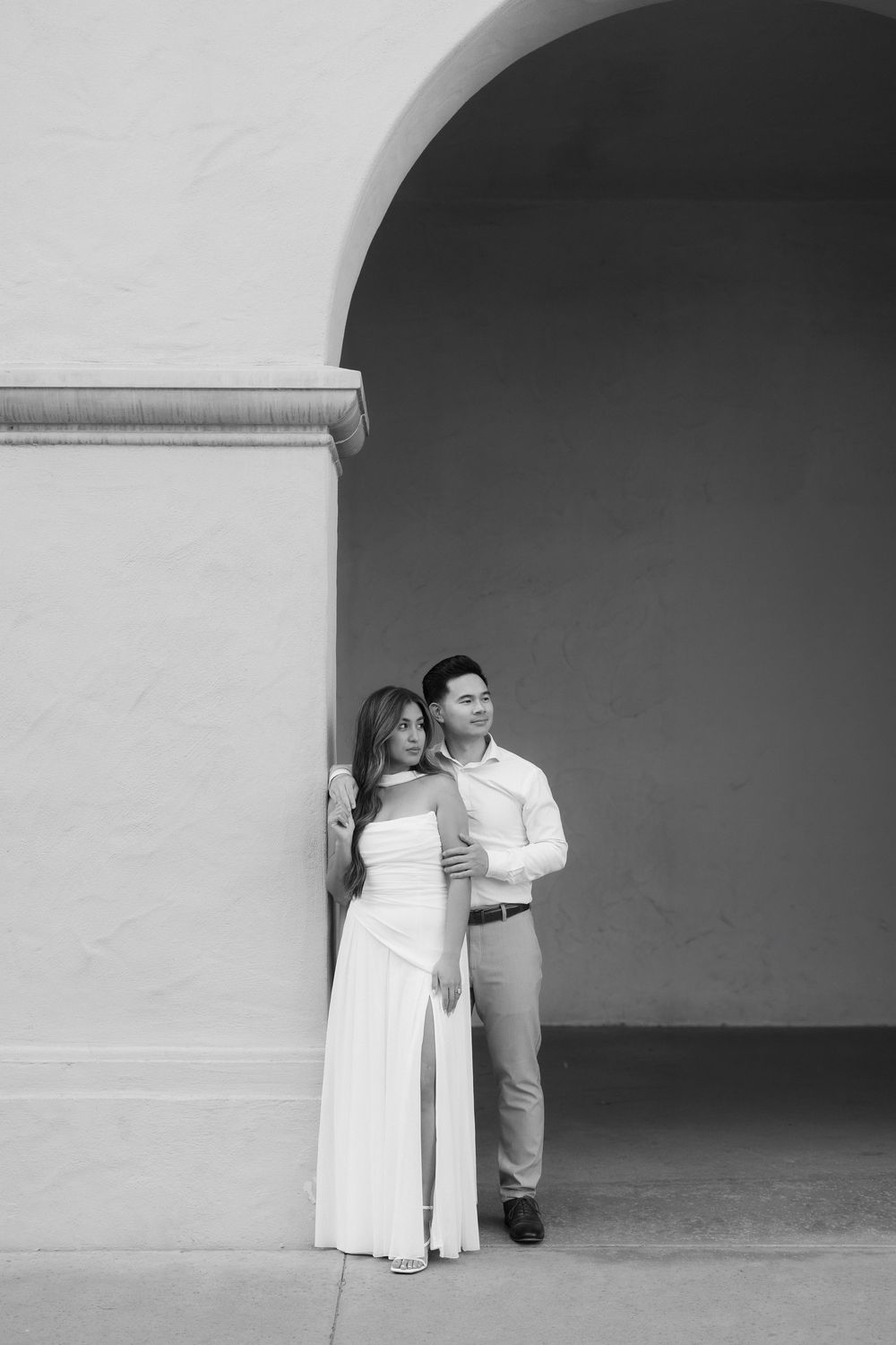 Black and white photo of a couple posing under an arch, woman in white gown leaning against wall with man behind her.